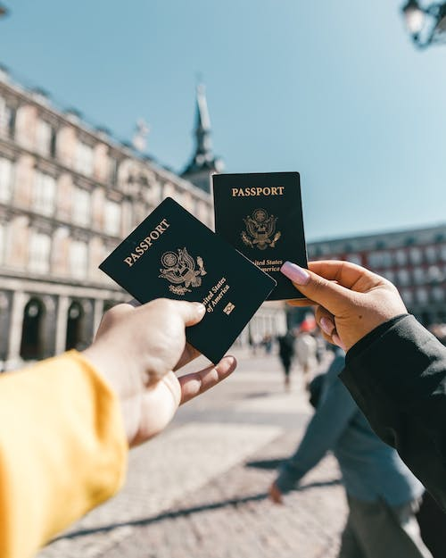 two people holding their passports on a busy cobbled street with a tall building in front of them.